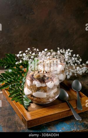 Classic Italian tiramisu layered dessert with cocoa powder and almonds in two glasses. Served on tray with spoons on old wooden table. Decorated white Stock Photo