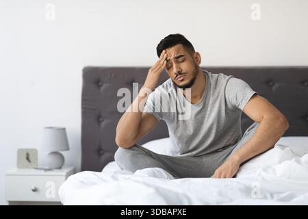 Tired Young Arab Man Sitting in Bed at Home Feeling Unwell With a Headache Stock Photo