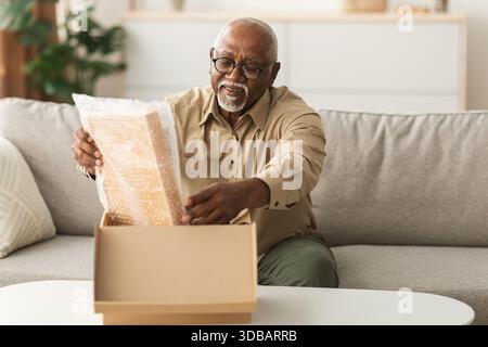 Senior Man Opening a Package at Home, Enjoying a Moment of Excitement and Curiosity While Seated on His Couch Stock Photo