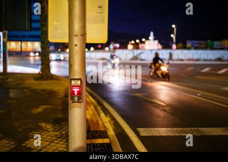 A close-up of a pedestrian crossing pole with a glowing red "PULSE EL BOTÓN ESPERE VERDE" (Press the Button Wait for Green) sign. In the blurred backg Stock Photo