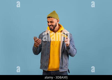 Happy Man Wearing a Green Beanie Looks at Phone While Holding a Credit Card in Front of a Blue Background Stock Photo