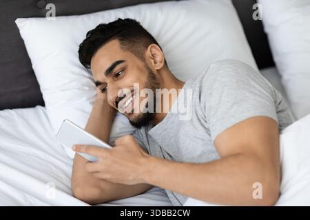 Handsome Arab Man Relaxing in Bed While Using Smartphone for Entertainment and Connection Stock Photo