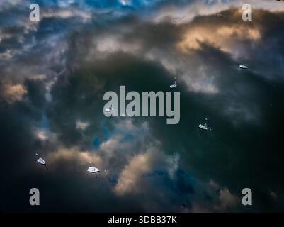 Aerial view of sailboats scattered on dark waters reflecting a dramatic sky with brooding clouds and streaks of golden light, Boston, Massachusetts, U Stock Photo