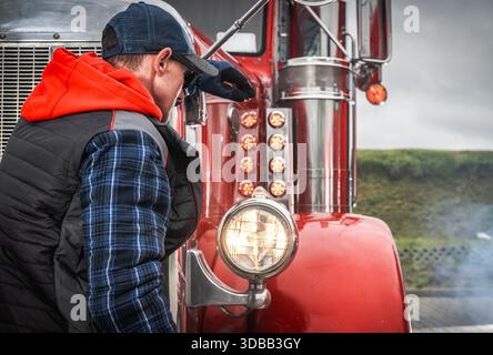A person stands beside a red truck looking at its front light while outdoors in a rural setting during the day. Stock Photo
