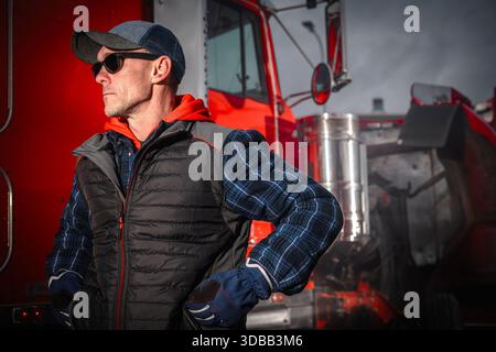 A man in a cap and sunglasses stands by a truck while wearing a jacket. He looks focused and relaxed in the moment. Stock Photo