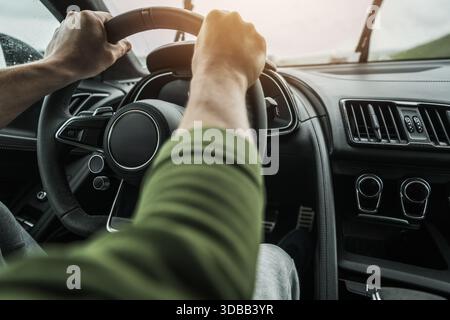 A person is holding the steering wheel of a car while driving on a wet road. Stock Photo