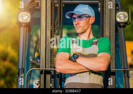 A worker in a green shirt and hat stands in a machine at a construction site. The sun shines on the scene. Stock Photo