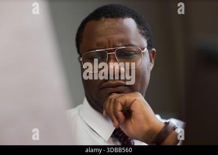 Middle aged Black man wearing glasses studying documents with thoughtful expression, hand resting on chin, office setting suggesting school principal reviewing paperwork Stock Photo