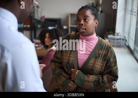 Young Black woman standing in office talking to middle aged Black man, another young Black woman sitting in background, school principal context suggesting administrative discussion Stock Photo