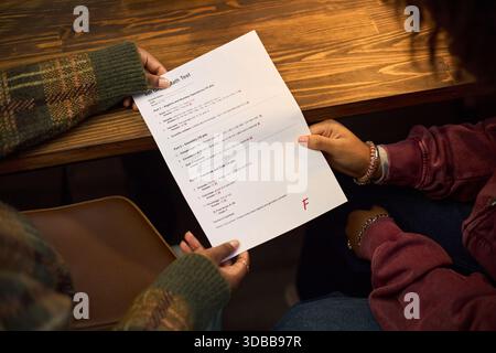 Two young adult women sitting at wooden table reviewing failed exam paper together, one woman holding test with visible red F grade, both focusing on academic results and discussing performance Stock Photo