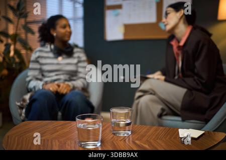 Middle aged Caucasian woman interviewing young Black woman in office setting, both seated and engaged in conversation, glasses of water and notepad on table in foreground Stock Photo