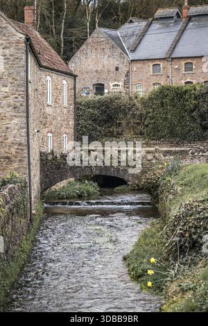 View of tranquil stream flowing under an ancient stone bridge, flanked by weathered brick buildings and lush greenery, Wells, England, United Kingdom. Stock Photo