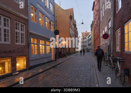 View of an old cobblestone street lined with colorful buildings glowing with warm light as people walk in the distance, Copenhagen, Denmark. Stock Photo