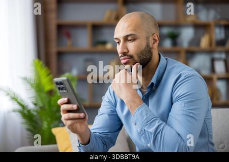 Young bald bearded man sitting on a sofa at home, focused on his smartphone while thoughtfully browsing and making a serious decision about content or information online Stock Photo