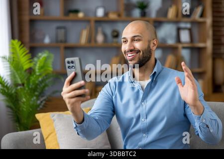 Young man relaxing on a sofa in a modern home, smiling and waving while engaged in an online video call using his smartphone, connecting with friends or family virtually Stock Photo
