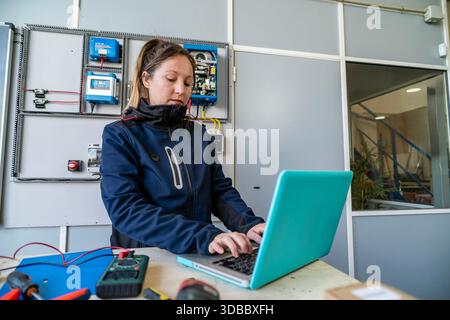 Female technician working on laptop at an electrical panel in an industrial workshop Stock Photo