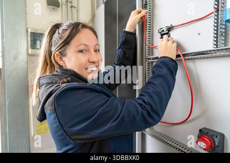 Woman technician working on an electrical switchboard, connecting red wires and smiling Stock Photo