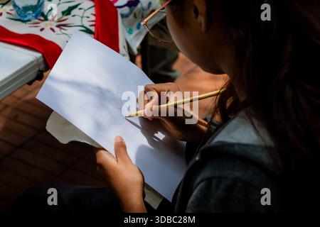 Profile view of a woman sitting outdoors, drawing a sketch with a pencil on white paper for an artisanal design. Stock Photo