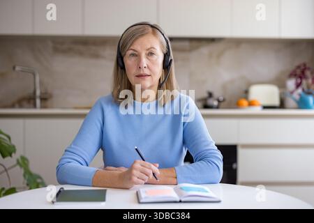 Mature woman in her kitchen wearing a headset and holding a pen, focused on a laptop during a video conference or e-learning session, taking notes and working from home Stock Photo