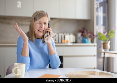 Senior woman feeling stressed and frustrated, having an angry phone call conversation while sitting at a kitchen table during the daytime, expressing complaints Stock Photo