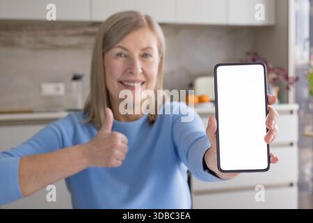 Senior woman smiling and holding a modern smartphone with a blank white screen while giving a thumbs up in approval, ideal for app or ad mockups and positive messaging Stock Photo