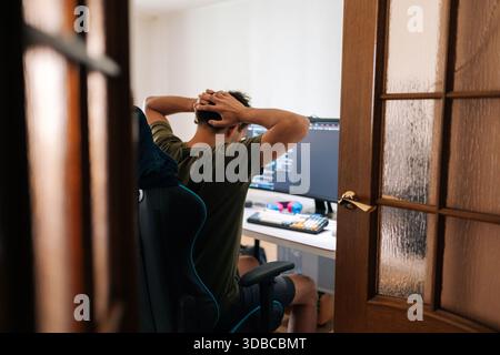 Back view of stressed programmer sitting in gaming chair in home office, holding head in hands after unsuccessful coding session, feeling overwhelmed Stock Photo