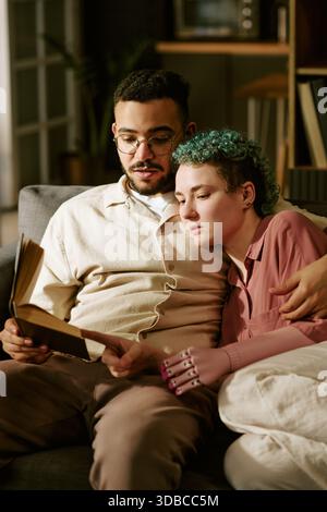 Young Adult Couple Reading Book Together on Sofa Embracing Stock Photo