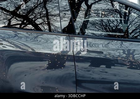 Close-up of a Tesla car door reflecting holiday lights and leafless winter tree branches in an urban setting Stock Photo