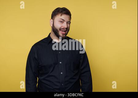 A man stands against a yellow background and looks at the camera. He smiles and winks. He wears a black shirt and has a beard. The setting is likely a Stock Photo