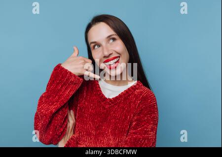 A woman shows a phone gesture with her hand and smiles brightly. She wears a red sweater and has long dark hair. The backdrop is blue and simple. Stock Photo
