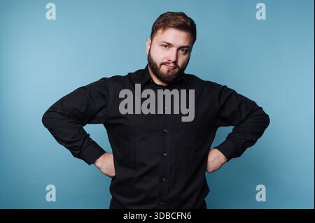 A man with a beard poses against a blue wall. He stands confidently with his hands placed on his hips. It appears to be an indoor setting with soft li Stock Photo