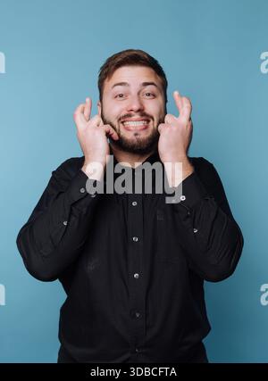 A man stands against a blue wall with a big smile, crossing his fingers in a gesture of hope. He wears a black shirt and looks cheerful and optimistic Stock Photo