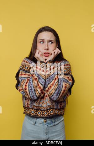 A woman stands against a bright yellow wall. She looks worried with her hands on her face. She wears a colorful patterned sweater and denim jeans. Her Stock Photo