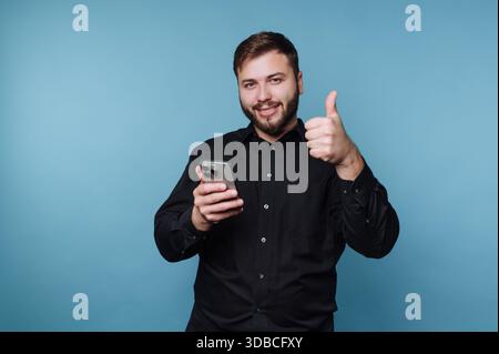 A man is smiling while holding a smartphone in one hand and giving a thumbs up with the other. He is in front of a plain blue wall. The scene is brigh Stock Photo