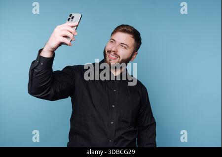 A man stands in front of a blue background. He holds a smartphone up and smiles while taking a selfie. He is wearing a black shirt and appears happy. Stock Photo