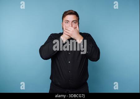 A man stands with wide eyes and covers his mouth with both hands. He appears shocked or surprised. The background is a plain blue, creating a clear fo Stock Photo