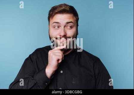 A man stands with a thoughtful expression, touching his chin with one hand. He has a light beard and is wearing a black shirt. The background is blue. Stock Photo