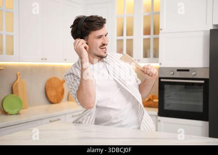Happy man in headphones with spatula singing at white marble table in kitchen Stock Photo