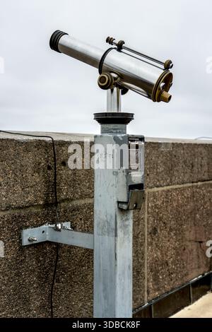 A metallic tourist telescope mounted on a stone wall overlooks a landscape under a cloudy sky Stock Photo