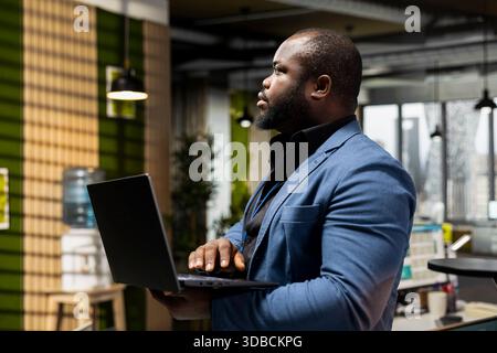 African american man focused on business analysis with laptop in hand, reviewing reports and charts using digital technology to enhance efficiency. Aligning resources to achieve greatness. Stock Photo