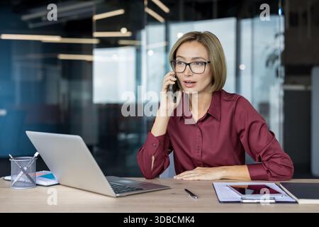 Asian businesswoman in modern office talking on cell phone while working on laptop at desk, focused on client communication, managing tasks and digital collaboration Stock Photo