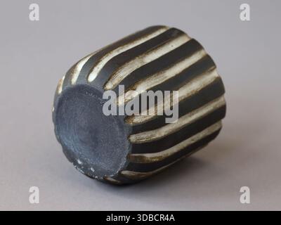 Studio shot of a small handmade black ceramic cup with carved striped texture. The pottery piece lies on its side against a neutral gray background. Stock Photo