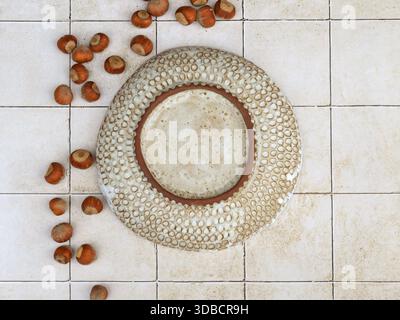 Top view of a handmade ceramic bowl with carved circular texture placed on tiled surface. Still life composition with hazelnuts arranged nearby, photo Stock Photo