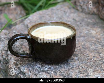 Minimalist still life of a handmade ceramic coffee cup filled with milk coffee, placed on natural stone outdoors. Dark glazed pottery, organic shape, Stock Photo