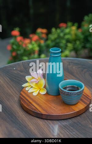 Black coffee served in ceramic cup with handmade bottle and tropical frangipani flowers on wooden board, outdoor natural light Stock Photo