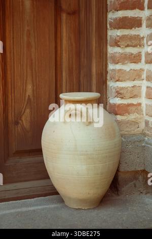 A large handmade clay jug placed beside a brick wall and a wooden door evokes the concept of simplicity and beauty. Stock Photo