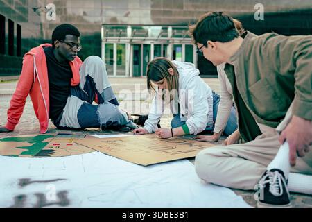 Group of young activists sitting on the ground and creating protest signs together outdoors, collaborating and preparing materials for a demonstration Stock Photo