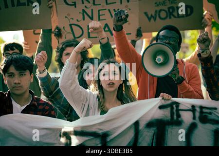 Group of diverse young activists protesting outdoors, raising fists, shouting slogans while holding banners and signs during a climate demonstration. Stock Photo