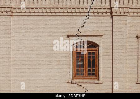Cracked brick wall with a wooden window, a deep jagged fissure symbolizing time's passage and structural fragility. Stock Photo