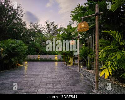 A tranquil evening scene in Bangkok features a stunning stamped concrete driveway surrounded by tropical greenery. Soft lights illuminate the vibrant plants as the sun sets on this peaceful retreat. Stock Photo